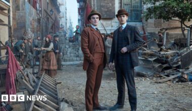 Young Sherlock stands with another man in an orange suit in a street made to look like a Victorian era London street. He wears a checked suit and a bowler hat