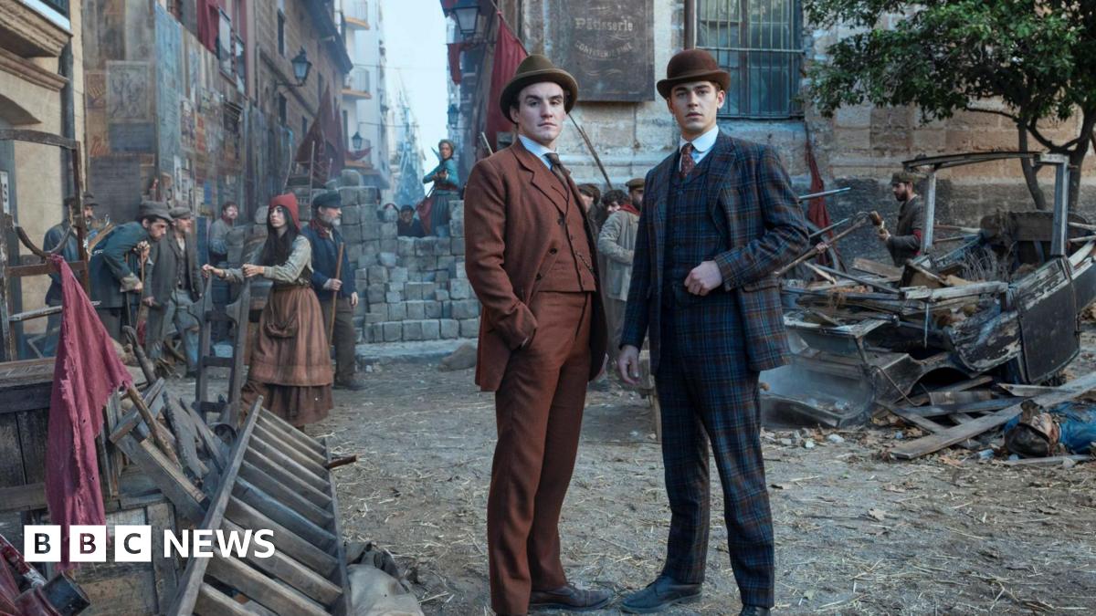Young Sherlock stands with another man in an orange suit in a street made to look like a Victorian era London street. He wears a checked suit and a bowler hat