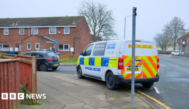 A blue and yellow marked police crime scene investigation van parked on the side of a road beside a row of terraced red brick houses. A black car is parked in front of it. The sky is grey.