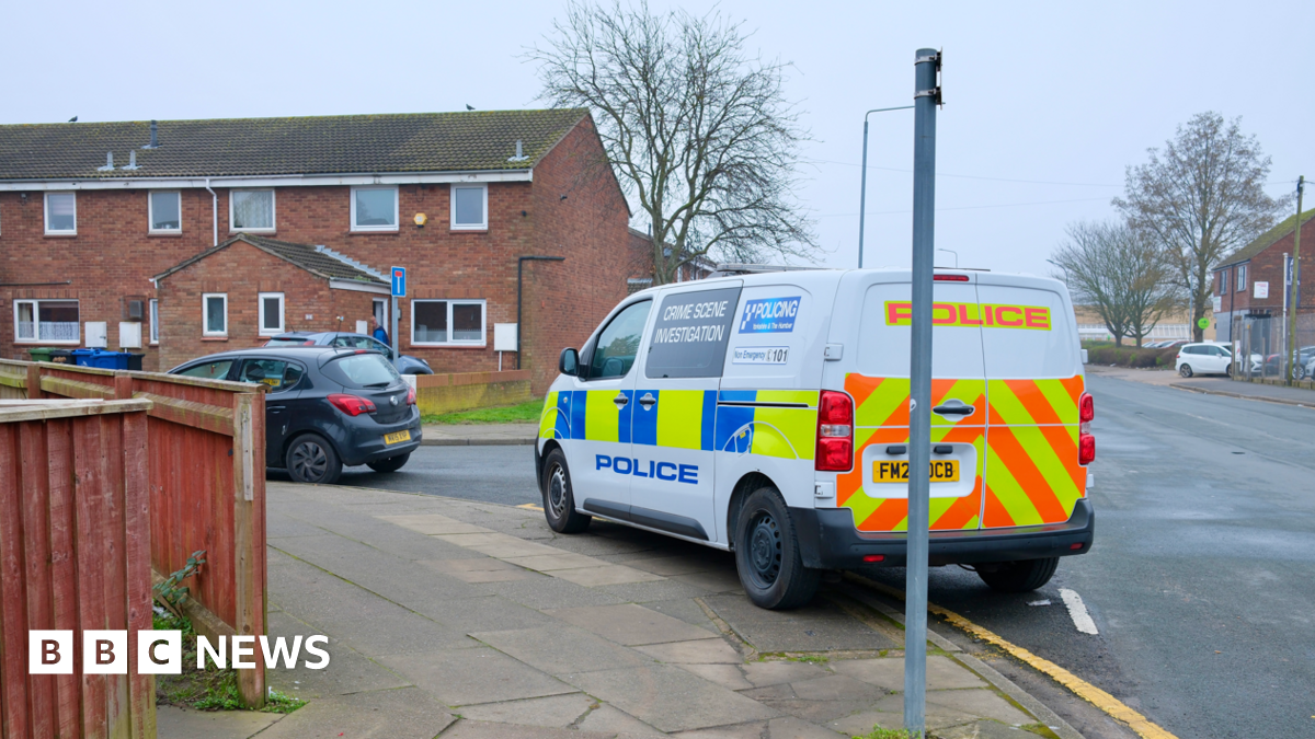 A blue and yellow marked police crime scene investigation van parked on the side of a road beside a row of terraced red brick houses. A black car is parked in front of it. The sky is grey.