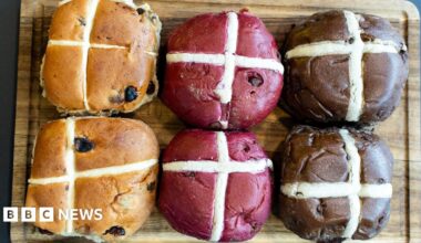 Creme eggs and other filled eggs sit among mini eggs, orange mini eggs and a partly unwrapped large Easter egg on a wooden board.