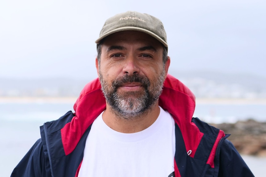 A close-up of a man with a greying beard, he is wearing a hat and rain jacket, the beach is behind him