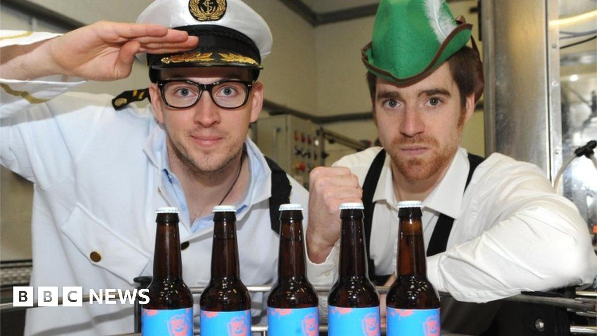 A man dressed as a sea captain next to a man wearing a green hat, posing for a photograph in front of bottles of beer