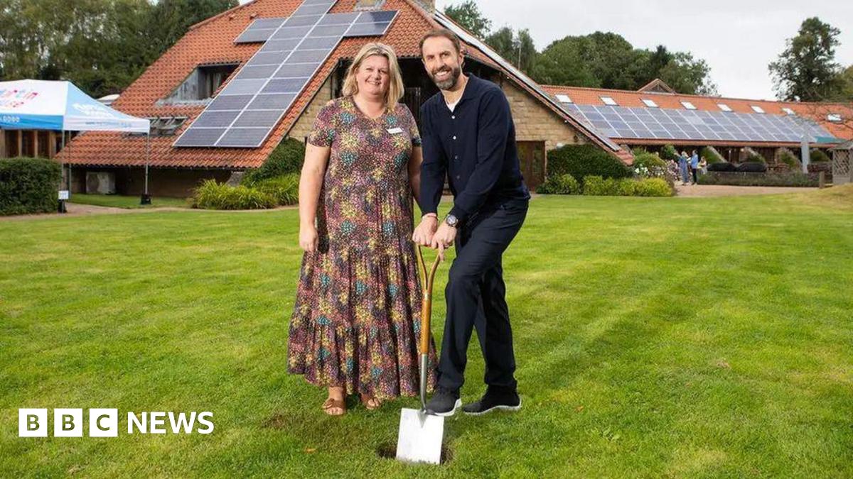 Two people stand on a neatly cut lawn in front of a modern building with a red‑tiled roof covered in solar panels. One of them holds a shovel, taking part in an event.