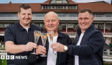 Paul pictured at a racecourse alongside his two sons, they all holding glasses of champagne and smiling. Paul is bald and stands in between his two sons who both have short dark hair. They are all wearing white t-shirts with navy blue jackets. Covered racecourse stands can be seen behind them.