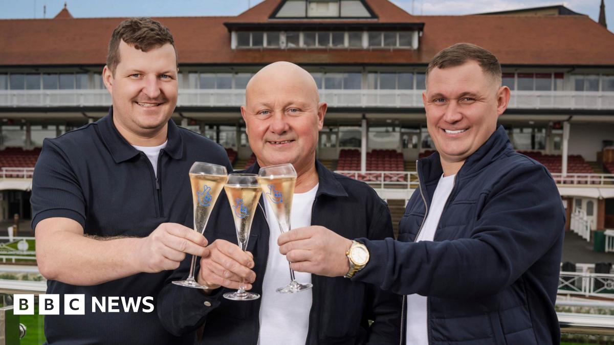 Paul pictured at a racecourse alongside his two sons, they all holding glasses of champagne and smiling. Paul is bald and stands in between his two sons who both have short dark hair. They are all wearing white t-shirts with navy blue jackets. Covered racecourse stands can be seen behind them.