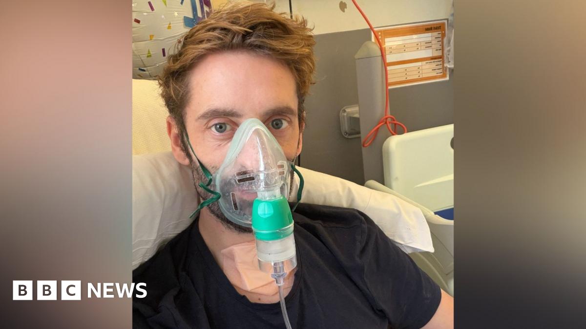 Daniel Harding wears an oxygen mask and looks into the camera. He has mid-length dark blond hair and is wearing a black T-shirt and is lying in a hospital bed.