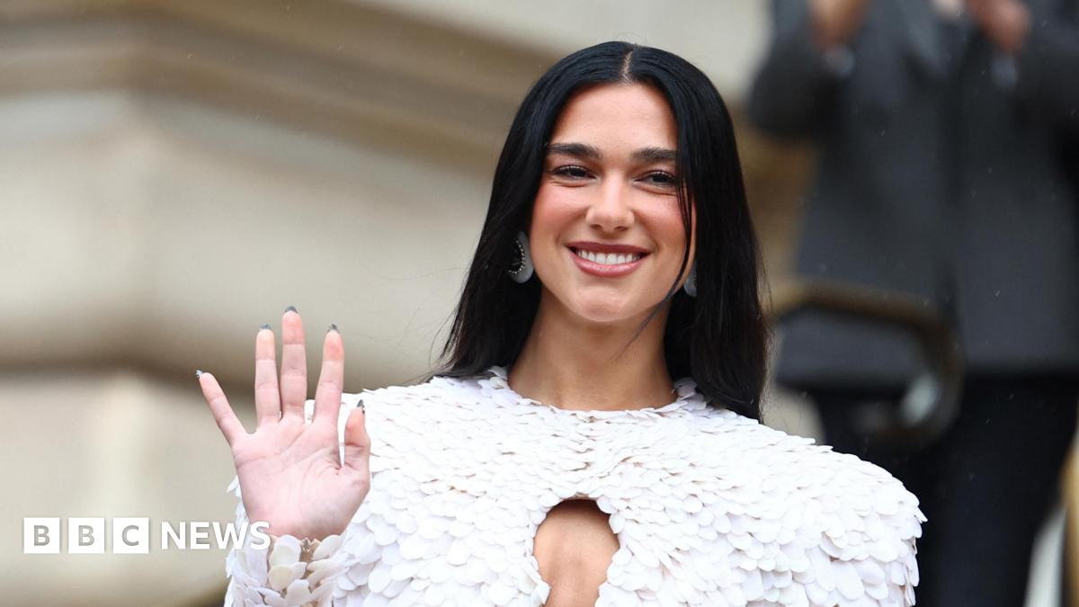 Dua Lipa wearing a feathered white outfit, waving at photographers and smiling.