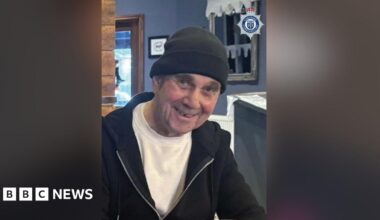 John McColl is seen smiling waring a black beanie hat, t-shirt and black hoodie.
