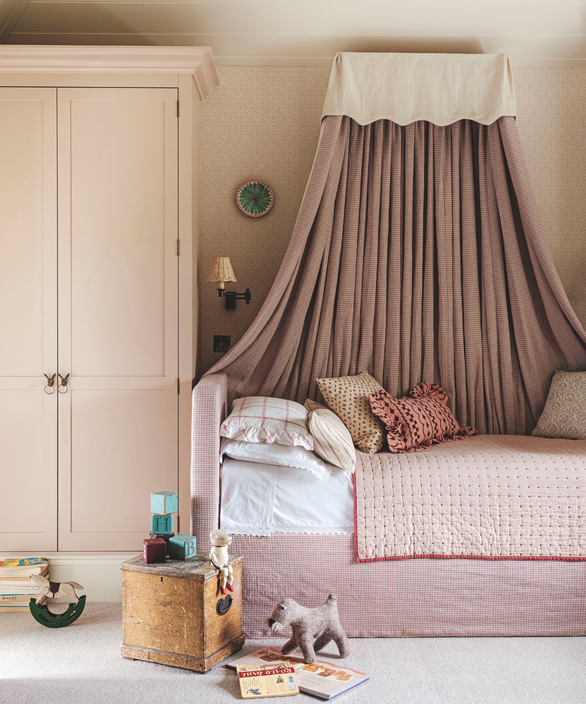 A charming child&rsquo;s bed nook featuring a draped pink-and-white checkered canopy, a matching pleated bed skirt, and a light pink wardrobe.