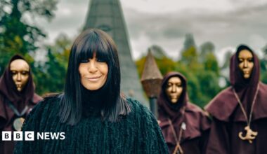 Claudia Winkleman wears a black coat as she smiles at the camera. Behind her is part of a castle and three people in black hoods and golden masks.