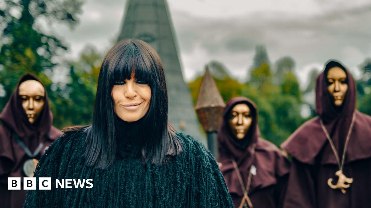 Claudia Winkleman wears a black coat as she smiles at the camera. Behind her is part of a castle and three people in black hoods and golden masks.