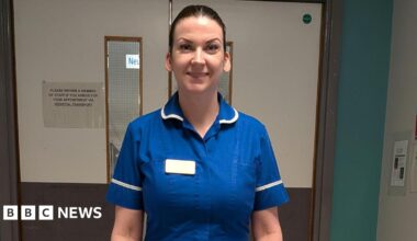 A smiling woman in a blue nurse's uniform with white trim standing in front of double doors in a hospital (Sheffield's Royal Hallamshire Hospital)