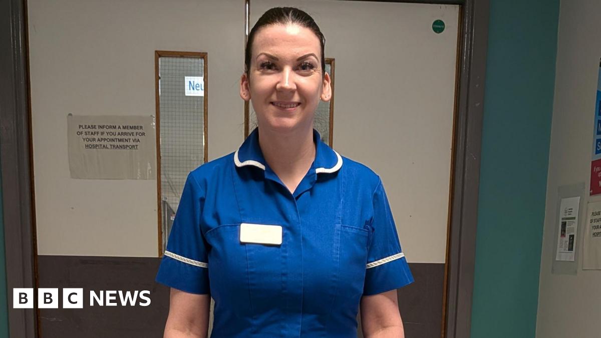 A smiling woman in a blue nurse's uniform with white trim standing in front of double doors in a hospital (Sheffield's Royal Hallamshire Hospital)