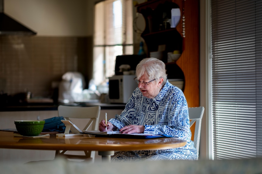 Suzenne, older woman with white hair, wearing a blue and white patterned top and glasses sitting at a table writing with a pen.