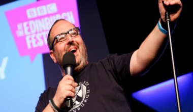 Justin Moorhouse on stage. He is holding a microphone and wearing a black t-shirt. He's wearing black glasses and smiling.