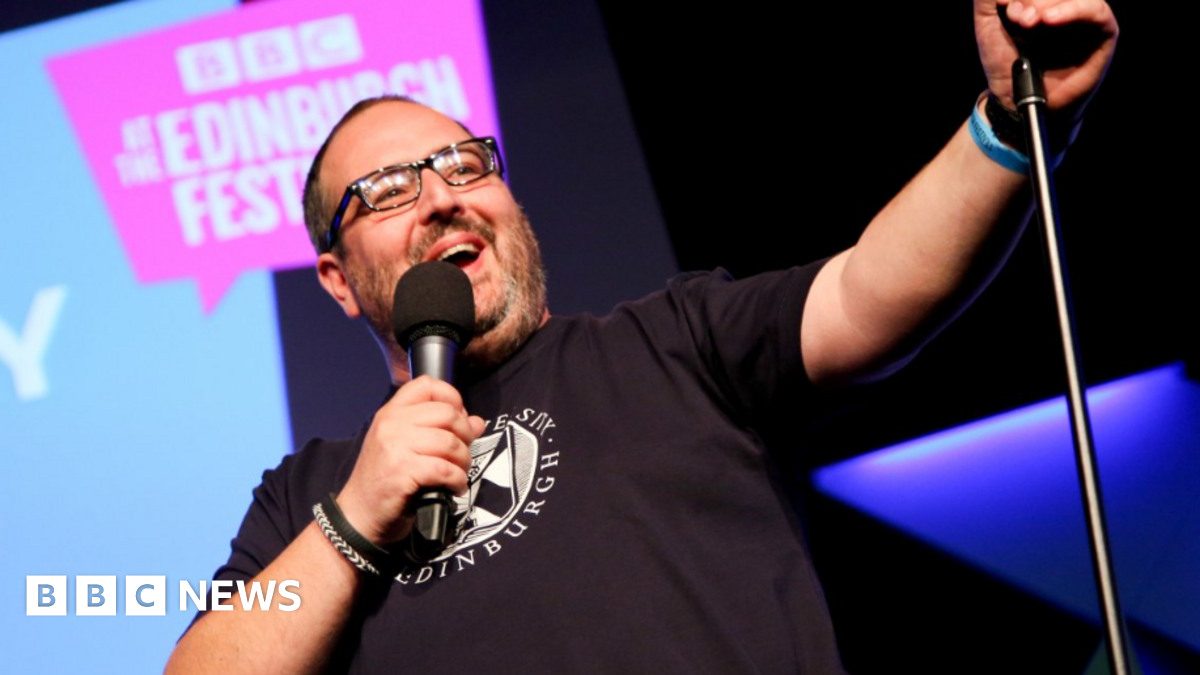 Justin Moorhouse on stage. He is holding a microphone and wearing a black t-shirt. He's wearing black glasses and smiling.