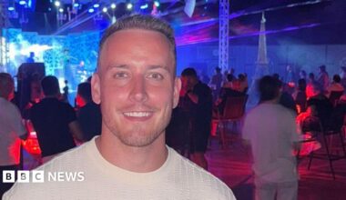 George, with short light brown hair and wearing a white T-shirt is smiling as he holds a drink with a red and white striped straw. He appears to be in a music venue, with musicians seen on stage behind him, along with a crowd of people. Red and blue light tints the rooms.
