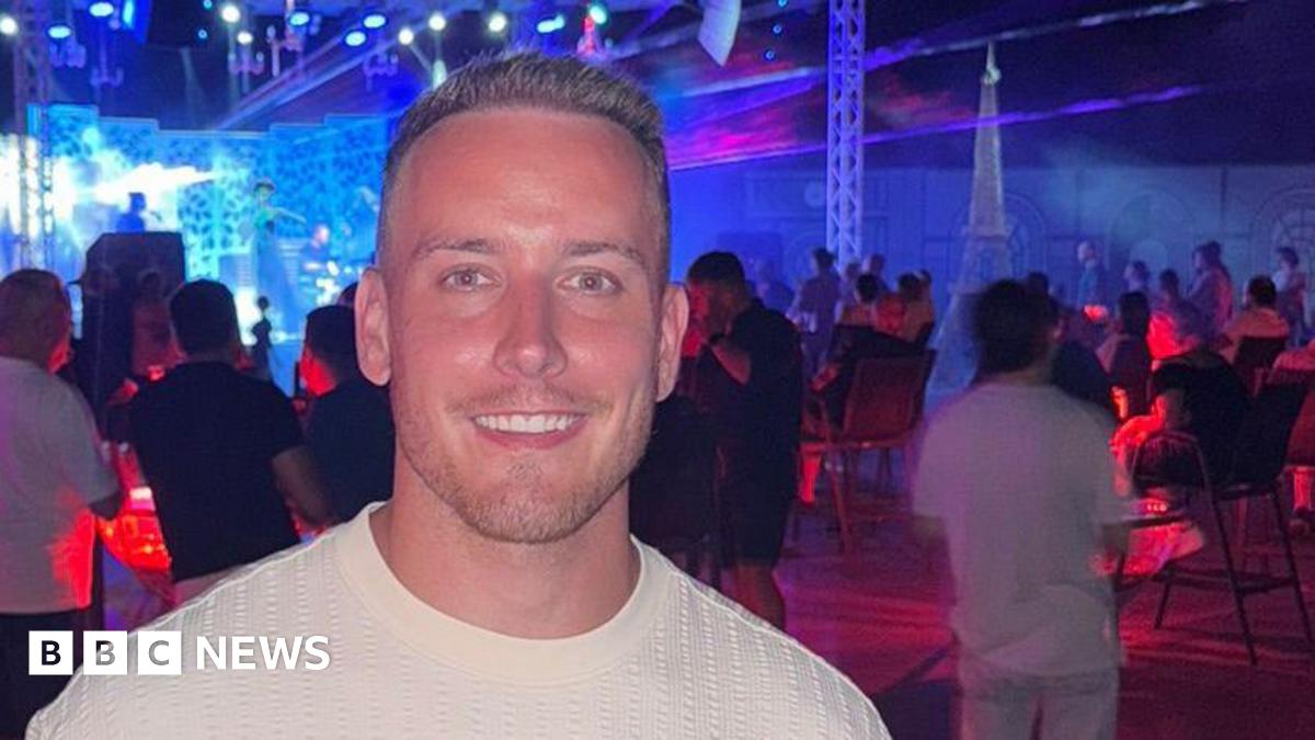 George, with short light brown hair and wearing a white T-shirt is smiling as he holds a drink with a red and white striped straw. He appears to be in a music venue, with musicians seen on stage behind him, along with a crowd of people. Red and blue light tints the rooms.