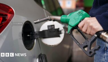 A man filling up a silver car with petrol. The pump handle is green and he wearing blue jeans and a jumper.