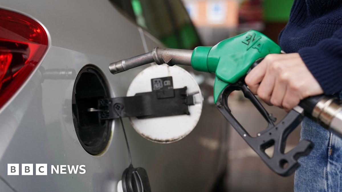 A man filling up a silver car with petrol. The pump handle is green and he wearing blue jeans and a jumper.