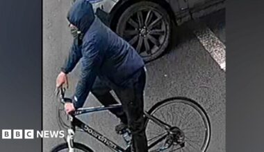 A man on a bike against a grey background. He is wearing a blue coat with the hood up and black trousers and is on a black bike with a white bar by the wheel.