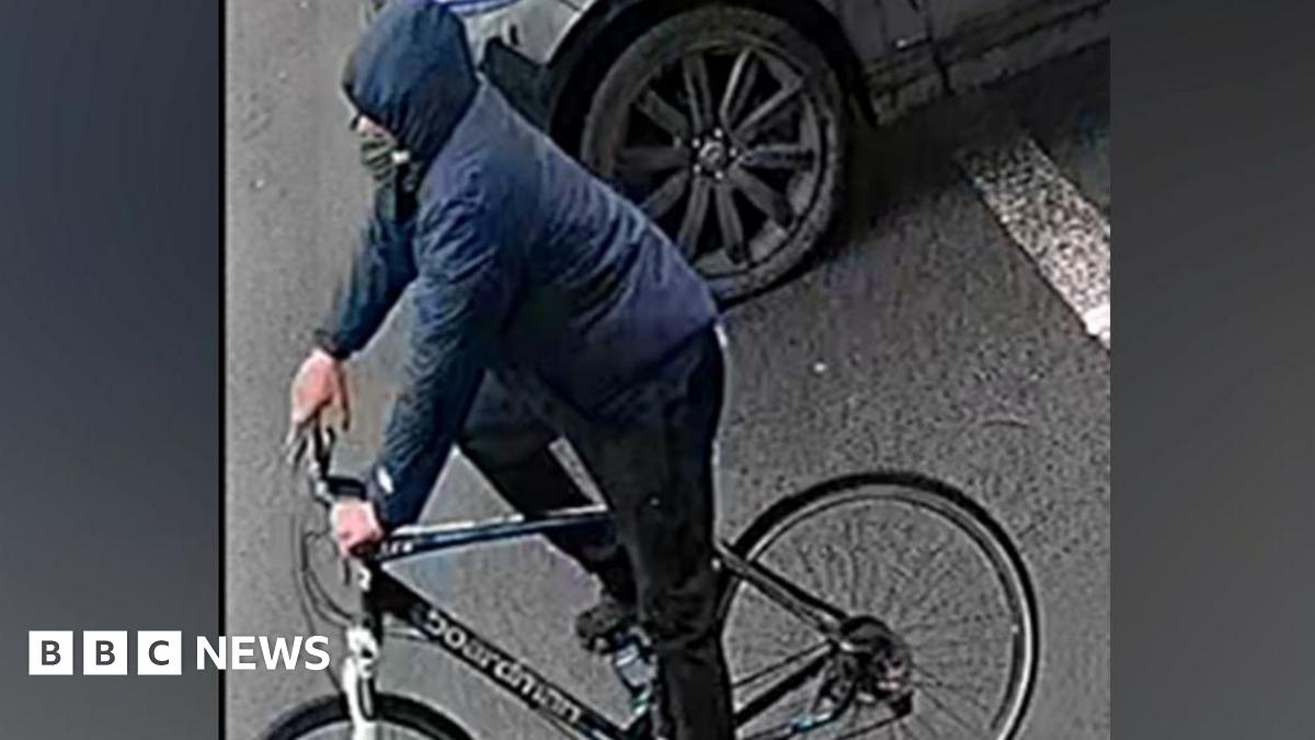 A man on a bike against a grey background. He is wearing a blue coat with the hood up and black trousers and is on a black bike with a white bar by the wheel.