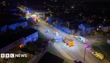 A nighttime aerial view shows a residential street filled with emergency vehicles with flashing blue lights. Several police cars, ambulances, and a fire engine are positioned along the road, which is cordoned off. Houses line both sides of the street, and the wider neighbourhood is visible in the background.