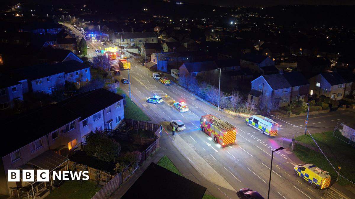 A nighttime aerial view shows a residential street filled with emergency vehicles with flashing blue lights. Several police cars, ambulances, and a fire engine are positioned along the road, which is cordoned off. Houses line both sides of the street, and the wider neighbourhood is visible in the background.