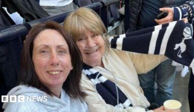 Two women sat in a football stadium wearing football scarves.