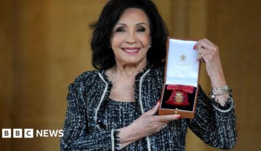 The image shows Dame Shirley Bassey holding her Companion of Honour award. She is wearing a black and white dress and matching jacket.
