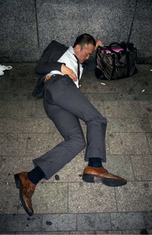A man in a suit lies on a city sidewalk with his eyes closed, one arm bent above his head, next to a brown bag. The ground is made of stone tiles and the background is a rough gray wall.