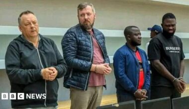 Four handcuffed men standing in front of a row of confiscated laptops.