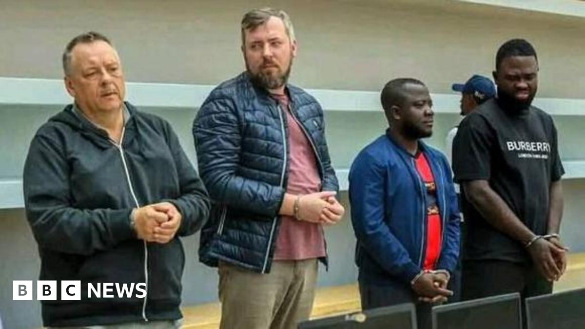 Four handcuffed men standing in front of a row of confiscated laptops.