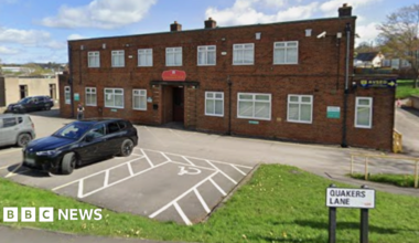 A two-storey brick built building stands surrounded by a car park and grassy area.
