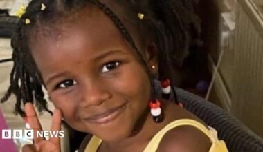 A woman with dark hair and a multiple colour jumper siting on a green sofa inside her living room.