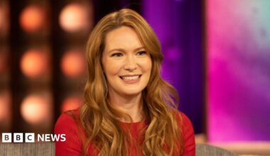 Picture of Sarah J Maas in a red top. She has ginger hair and is sat in front of a blurred background