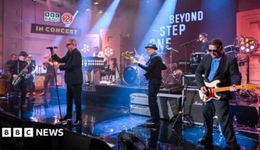 The band Madness perform on stage at a BBC Radio 2 concert. One man is holding a microphone while the others are playing various instruments.