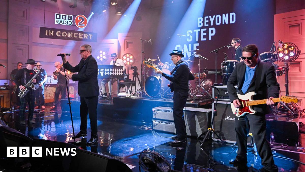 The band Madness perform on stage at a BBC Radio 2 concert. One man is holding a microphone while the others are playing various instruments.