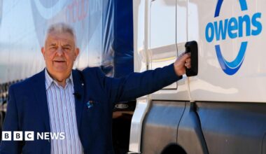 Huw Owen, wearing a blue blazer and striped shirt, stands next to the cab of one of his branded Owens Group lorries. He is holding the door handle and smiling at the camera on a sunny day.