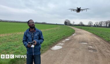 Dr Emmanuel Zuza stands on the track in the middle of a young wheat field and uses a console to fly a white drone that hovers just above head height