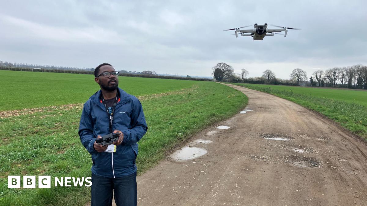 Dr Emmanuel Zuza stands on the track in the middle of a young wheat field and uses a console to fly a white drone that hovers just above head height