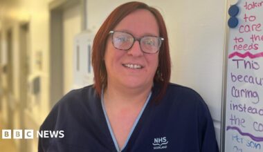 Nurse Katie Anderson smiling at the camera. She's wearing blue scrubs and has short red hair and glasses. She is standing next to a whiteboard with notes.