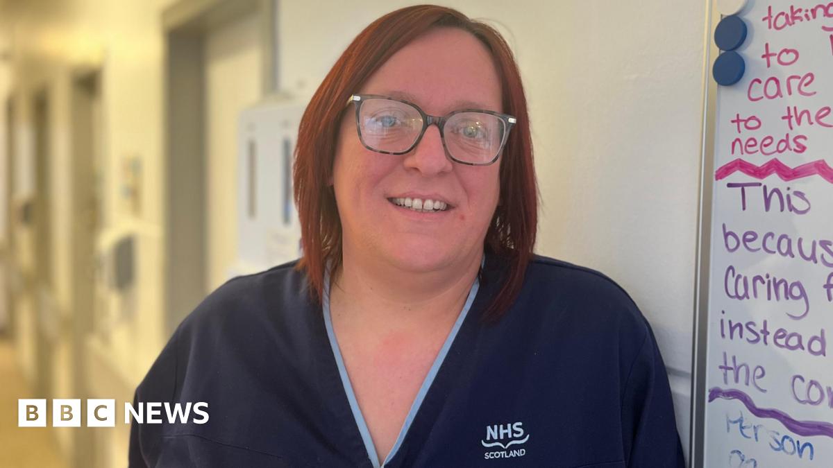 Nurse Katie Anderson smiling at the camera. She's wearing blue scrubs and has short red hair and glasses. She is standing next to a whiteboard with notes.