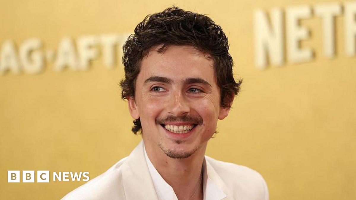 Timothée Chalamet smiling. He is wearing a white shirt and suit jacket. He has dark brown hair. The background behind him is gold.