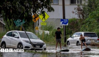 Two teenagers on surf  boards in shallow flood waters on a street next to a disabled grey car.