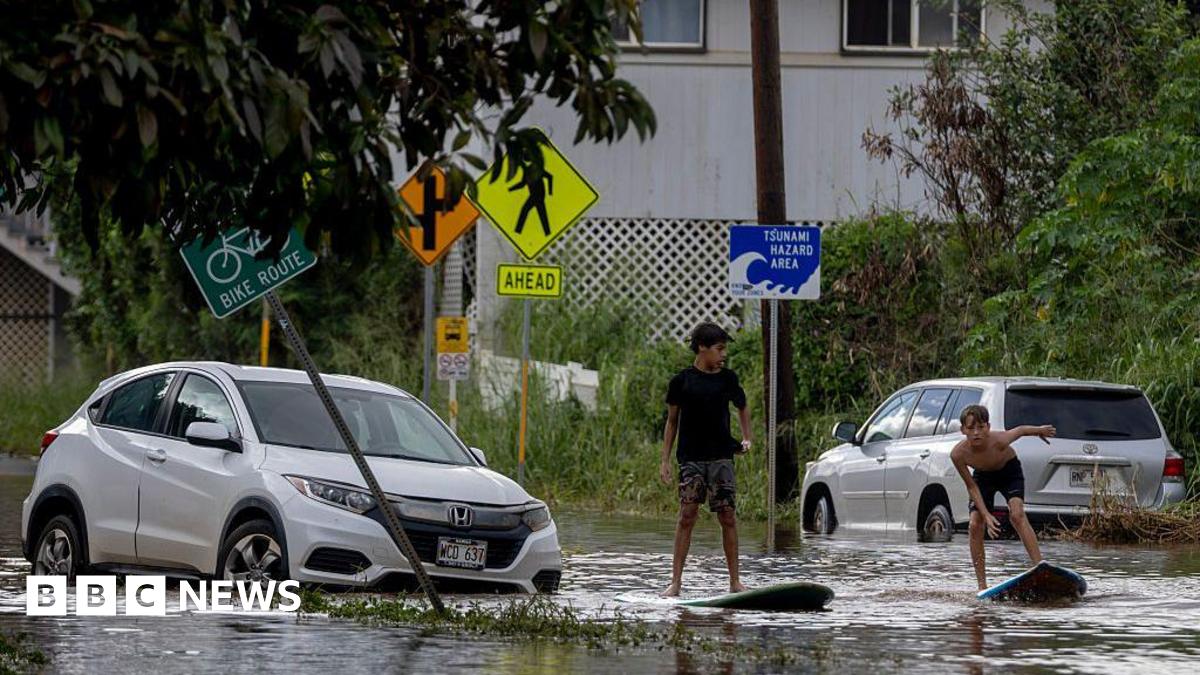 Two teenagers on surf  boards in shallow flood waters on a street next to a disabled grey car.