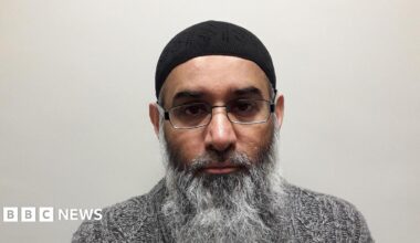 Mugshot of Casciani who has a grey bear and is staring at the camera, wearing a grey jumper, metal glasses, and  a black kufi prayer cap