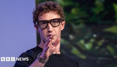 Mark Zuckerberg wearing thick black rimmed smart glasses and gesturing with his hand.