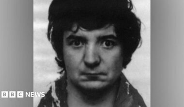 A black-and-white police mugshot of Peter Sullivan, taken in 1986. In the photograph he has black bushy hair and is wearing a woolly coat.